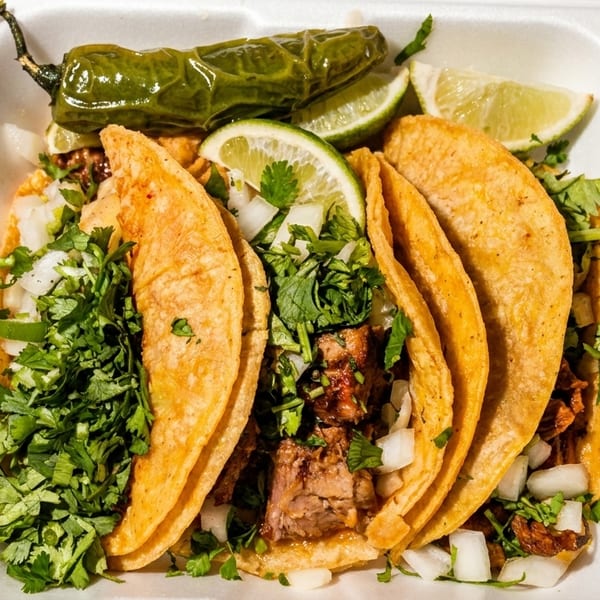 Four street tacos with meat, cilantro, and onions, served with lime wedges and a grilled jalapeño.