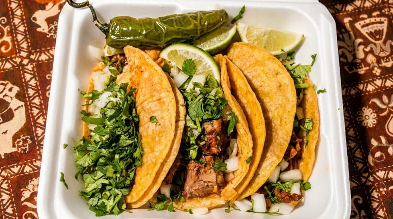 Four street tacos with meat, cilantro, and onions, served with lime wedges and a grilled jalapeño.