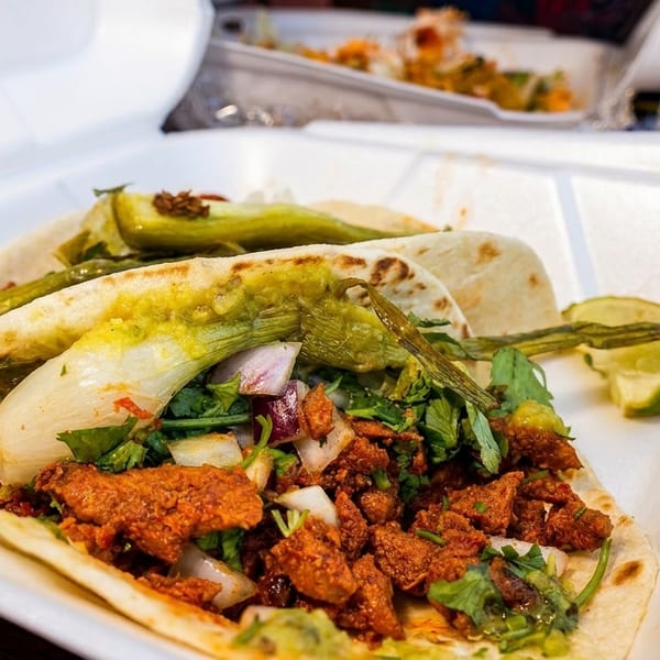 Two vibrant tacos with meat, cilantro, onions, and green chiles in a white takeout container.