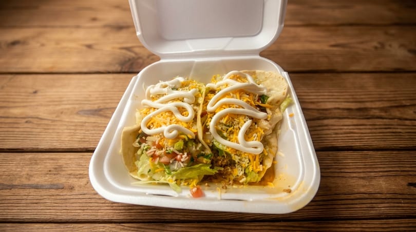 Two tacos with lettuce, cheese, and sour cream in a white takeout container on a wooden table.